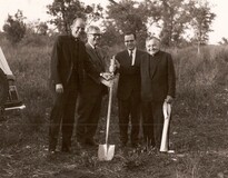 Sepia photo of four men in dark suits and clerical dress standing in an overgrown field with trees behind them, holding shovels for the groundbreaking.