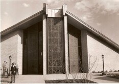 Sepia photo of the front of the original St. Theresa church: a steep peaked roof, a tall latticed window between two pillars, lamp posts on either side, and a few people out front.
