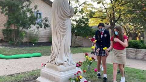 Students in school uniforms and face masks leave flowers at the foot of a white statue outside.