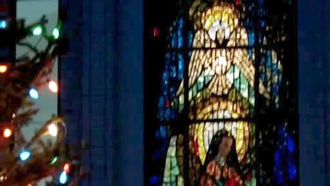 A lit Christmas tree beside a stained-glass window of a saint.
