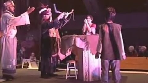 Two kings in robes raise their arms on stage, while a woman behind a table and a boy in a vest look on.