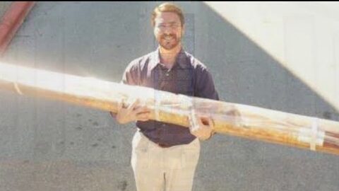 A smiling man holds a long organ pipe, wrapped in plastic and tape.
